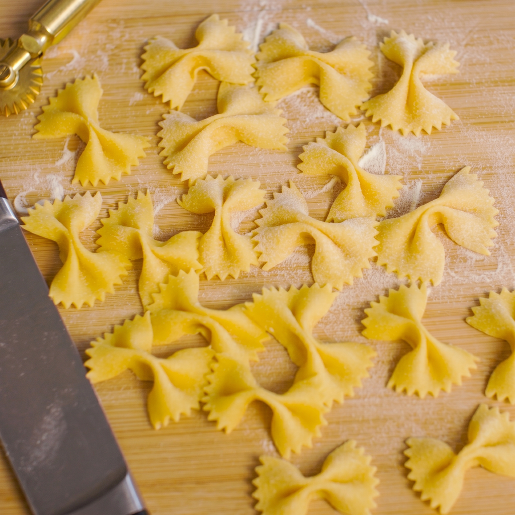 Uncooked bowtie pasta pieces scattered on a wooden cutting board with a metal rolling pin visible in the lower left corner.