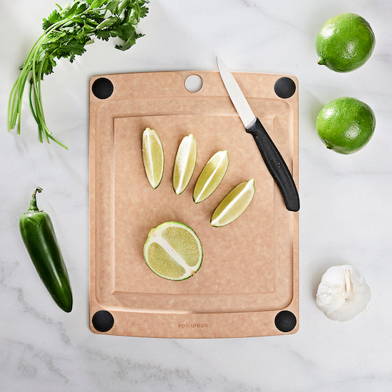 Wood composite Epicurean all-in-one series natural cutting board with sliced limes, a knife, and vegetables on a marble surface