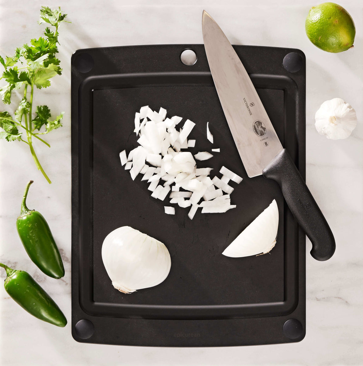 Black Epicurean non-slip cutting board with chopped onions, a knife, and vegetables on a white surface