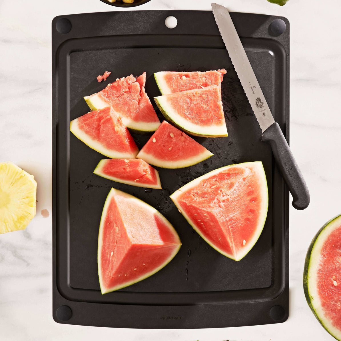 Sliced watermelon on a black Epicurean all-in-one series cutting board with a knife, on a white marble surface.