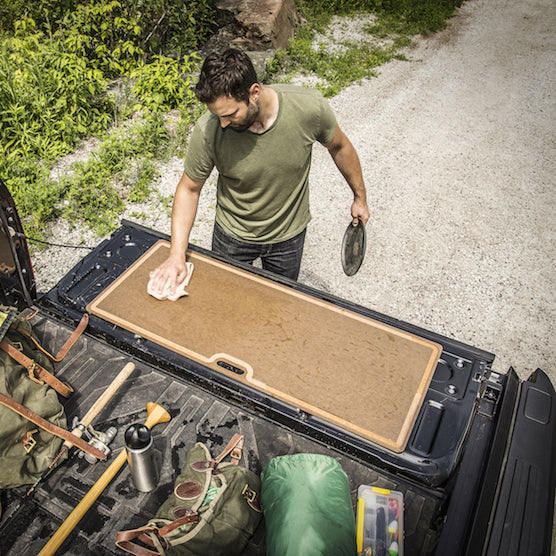 Man cleaning a Epicurean big game and butcher board in the back of a truck with outdoor scenery.