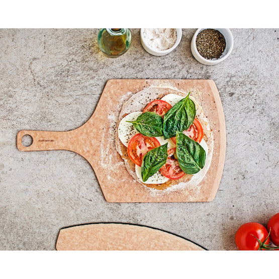 Epicurean pizza peel with pizza dough topped with tomatoes and spinach on a concrete surface.