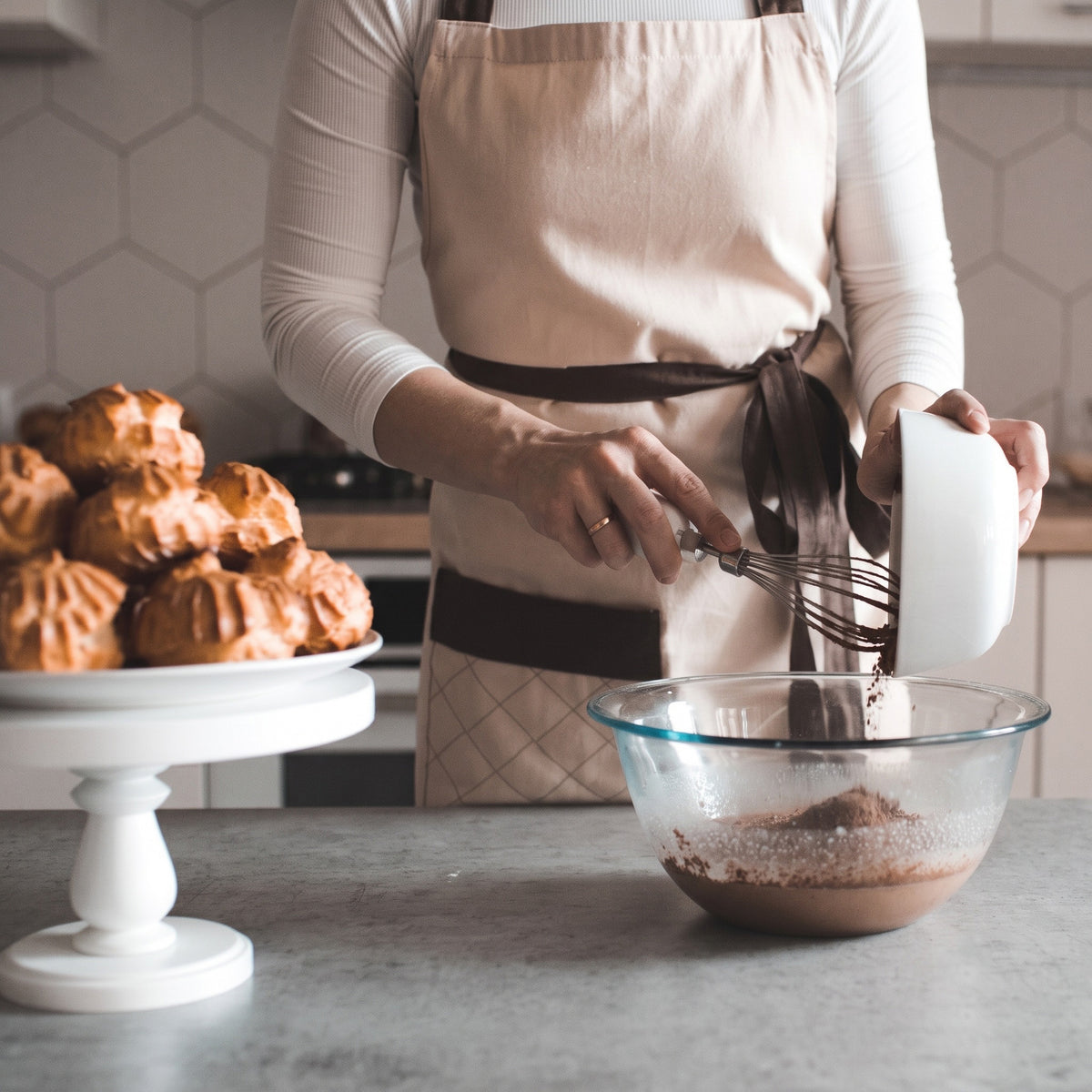 A person wearing a beige apron with dark brown trim mixes ingredients in a glass bowl on a kitchen counter, with baked pastries on a white cake stand in the background.