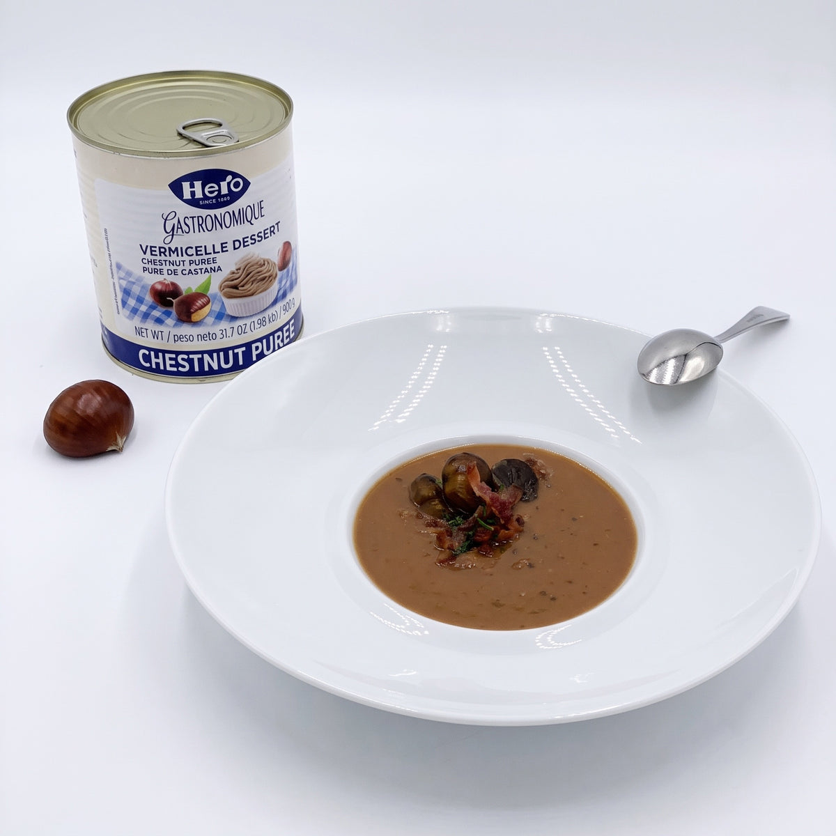 A can of Hero Gastronomique Chestnut Puree dessert next to a white bowl filled with the smooth brown chestnut soup, garnished with a few chestnuts and dried fruit pieces, with a small spoon on a white background.