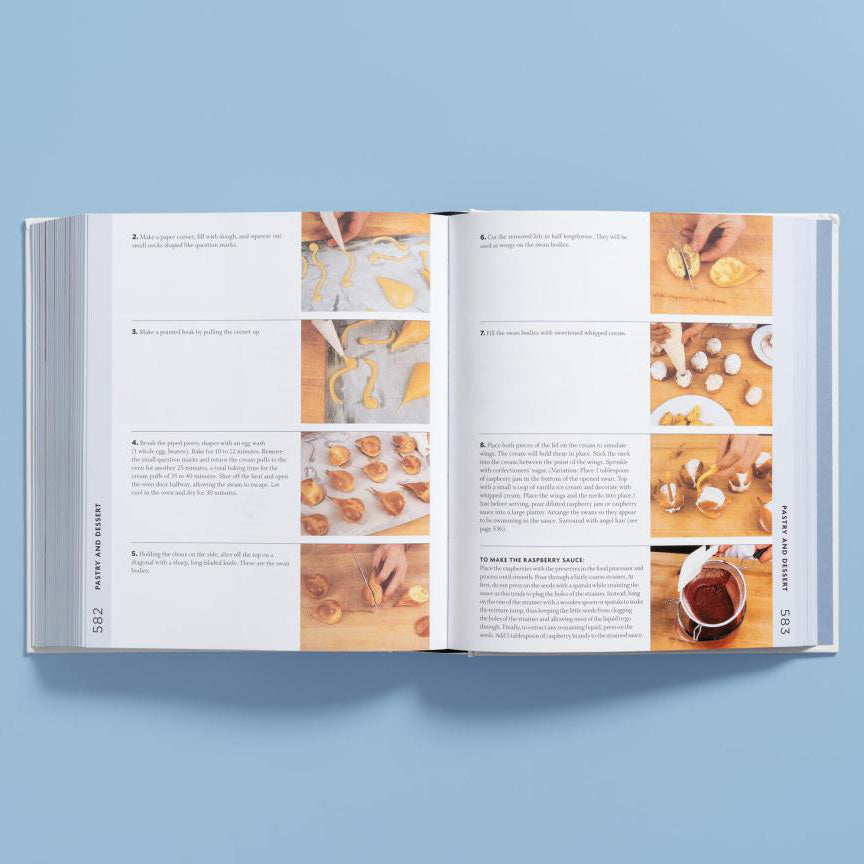 An open cookbook spread showing recipe pages with text and multiple small food preparation photos, photographed against a light blue background.