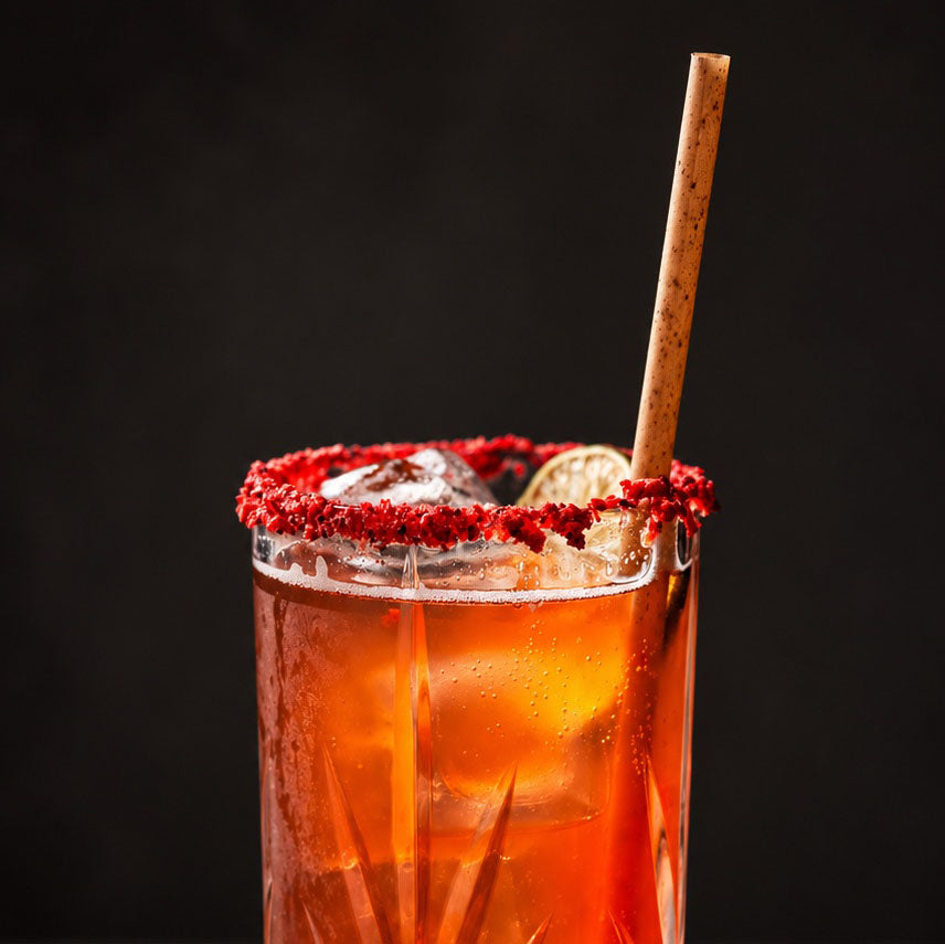 A close-up of a highball glass filled with an amber-orange cocktail, rimmed with strawberry pieces, containing ice and a wooden straw, set against a dark background.