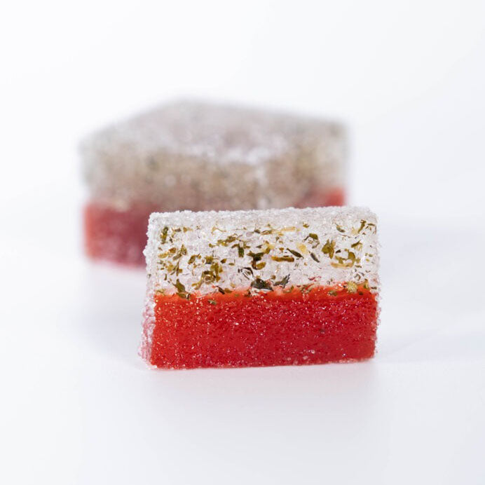 Two square layered sweets on a white background, with one in sharp focus in the foreground and one blurred in the background. The front sweet has a red bottom layer and a white top layer with dried herbs, while the back sweet shows a gray-brown top layer and a red bottom layer.