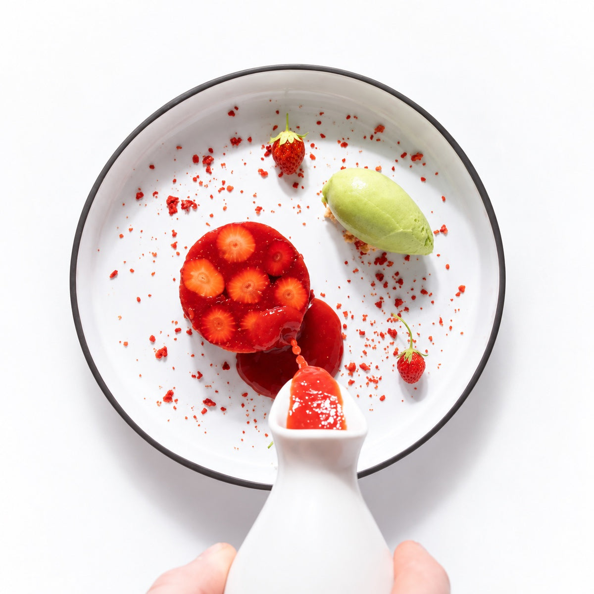 A top-down view of a white plate with a black rim, containing a strawberry dessert, a green ice cream, and scattered strawberry pieces, with a hand holding a small white bottle pouring more strawberry sauce onto the plate against a white background.