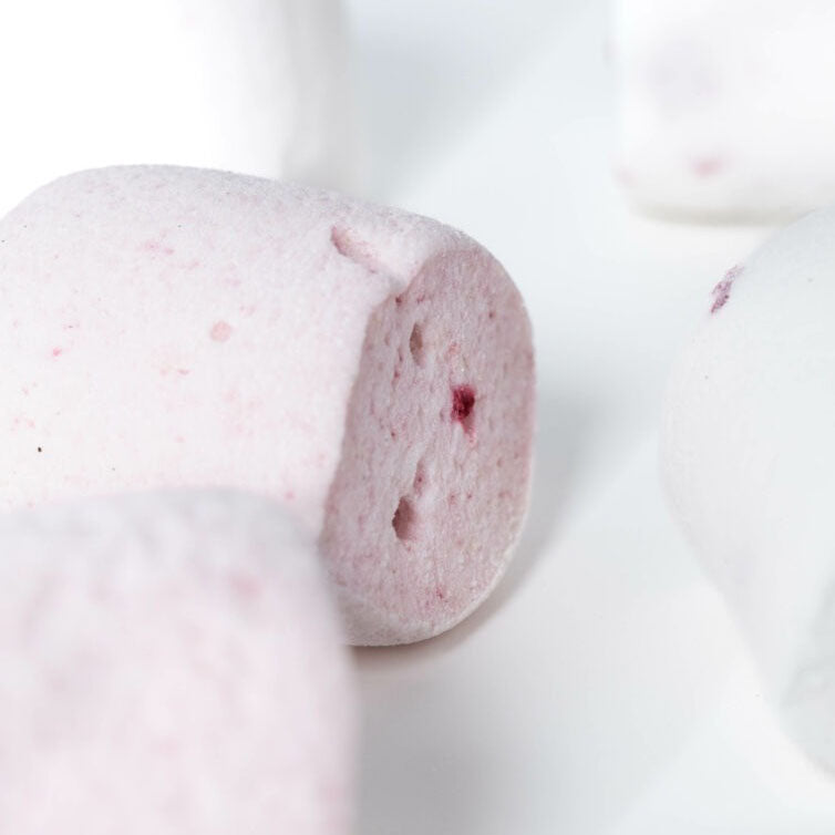 Close-up of a pale pink marshmallow with visible pink raspberry pieces embedded inside, photographed on a clean white background with other marshmallows softly blurred.