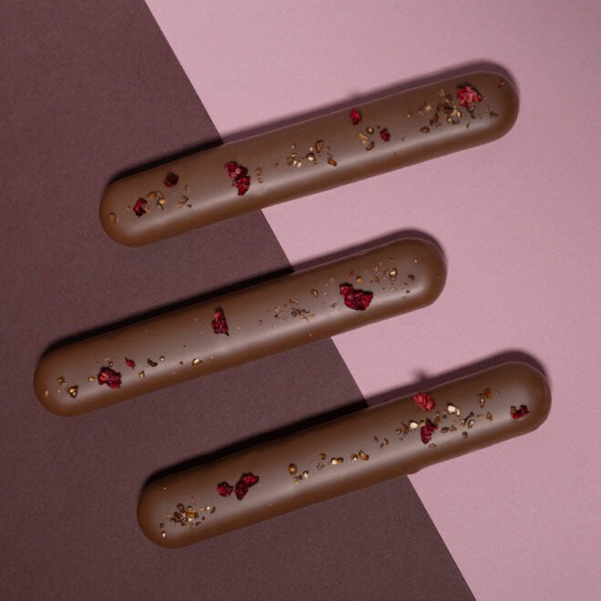Three chocolate-covered stick-like confections with scattered red dried raspberry pieces and small gold flakes, arranged diagonally on a split brown and pink background.