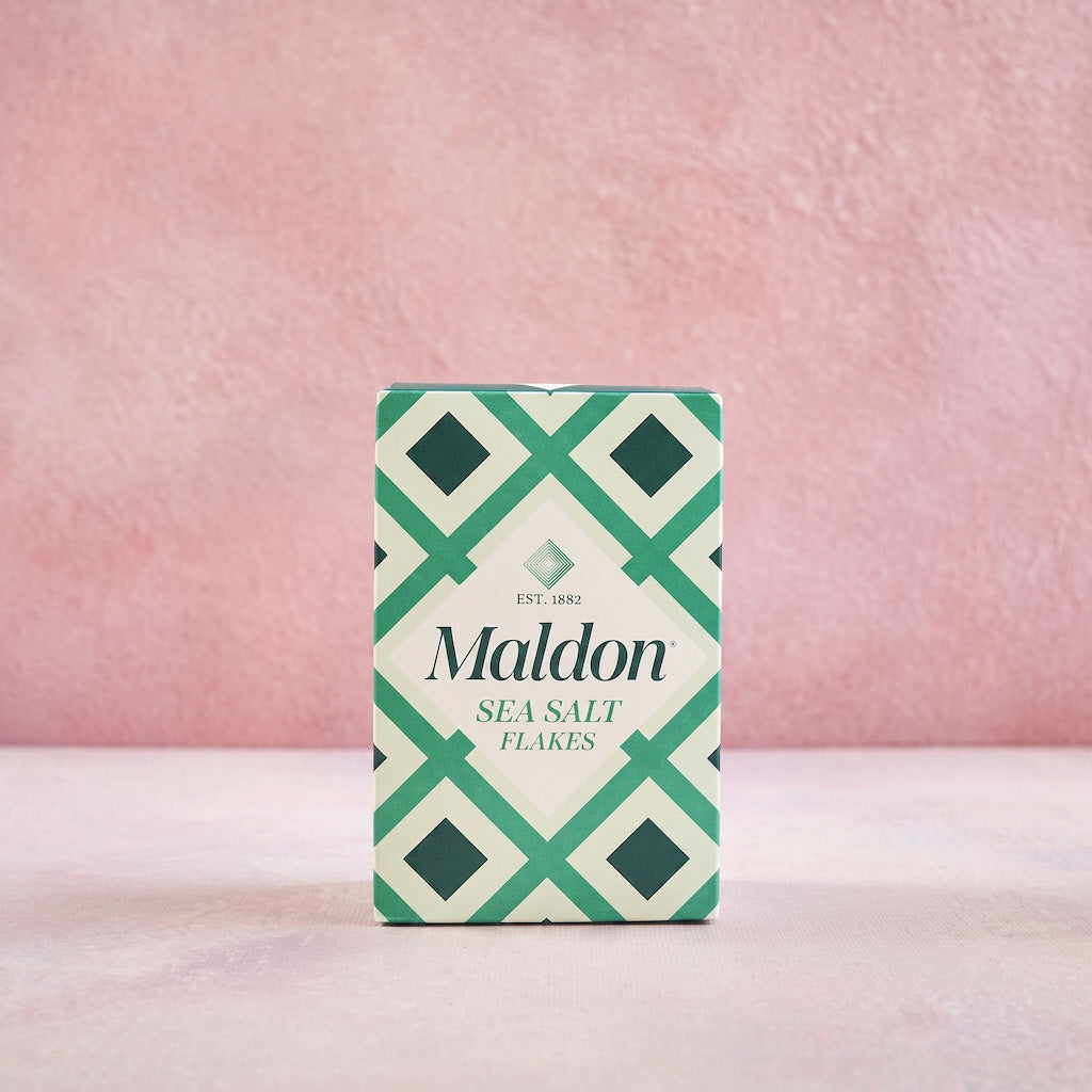A box of Maldon Sea Salt Flakes standing upright on a light pink surface against a textured pink background. The packaging features a green and white geometric pattern with the brand name 'Maldon' in black text.