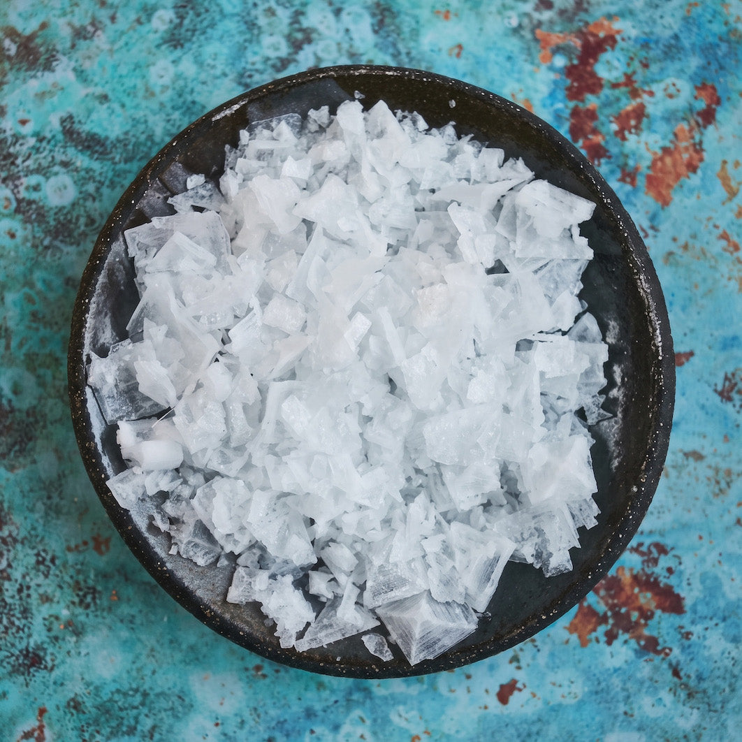 Coarse white crystal Maldon salt flakes piled in a round dark bowl, set against a textured turquoise and rust background.