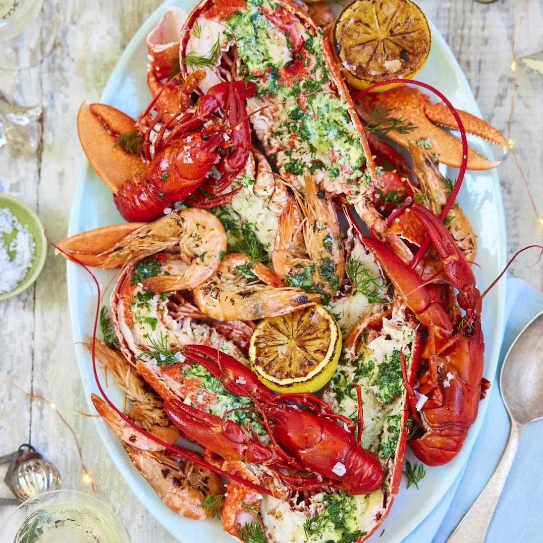 A large seafood platter on a light blue oval plate, featuring cooked lobster tails, red lobsters, shrimp, melted cheese, chopped green herbs, and lemon slices, photographed from above on a rustic white wooden table with a spoon and small garnish bowls nearby.
