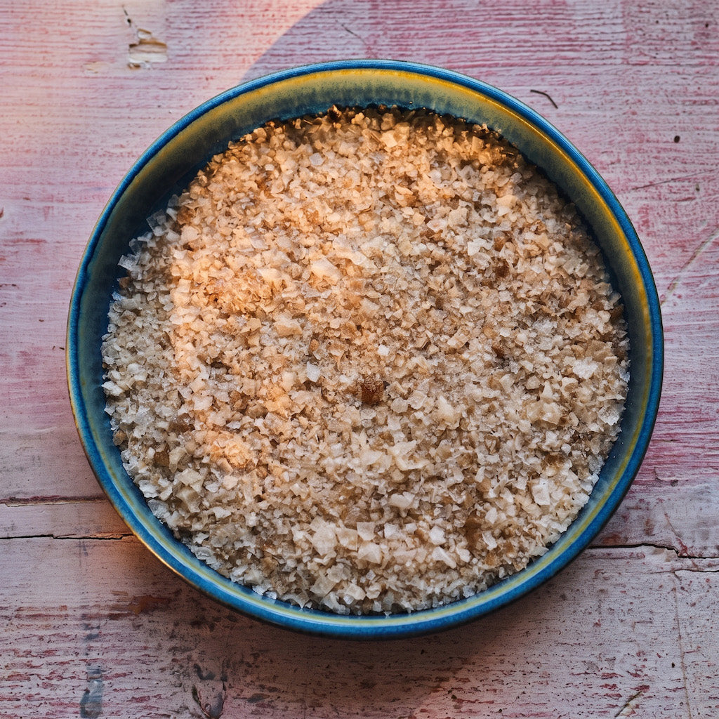 A top-down view of coarse Maldon smoked sea salt or mineral salt granules in a round blue ceramic bowl, set on a rustic, weathered pinkish-red wooden surface.