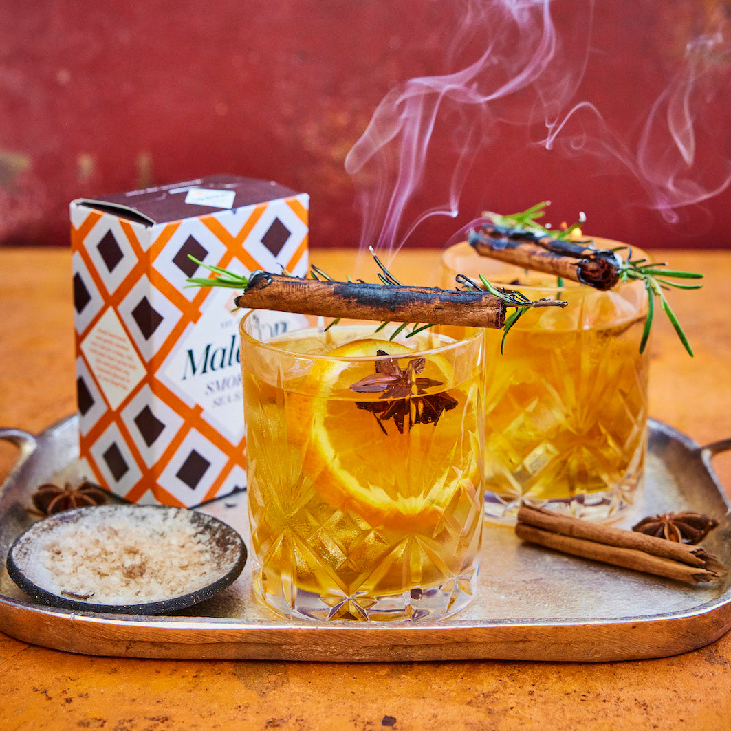 Two clear glasses filled with whisky, garnished with Maldon smoked sea salt, orange slices, star anise, cinnamon sticks, and rosemary sprigs, set on a metal tray beside a patterned box and a small dish of spices. Smoke rises from the glasses against a rustic red-brown background.