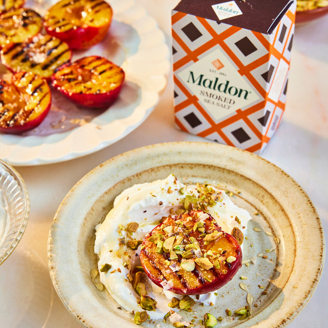 A box of Maldon Smoked Sea Salt sits on a table next to a plate of grilled plums and a dessert topped with nuts and sea salt, in a bright, clean setting.