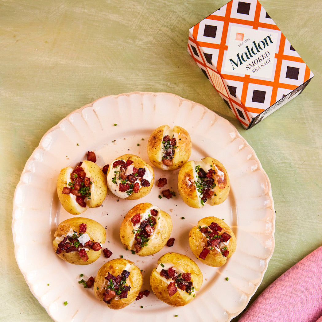 A plate of baked potato halves topped with bacon and herb cream, with a box of The Maldon Smoked Salt in the corner on a textured green surface.