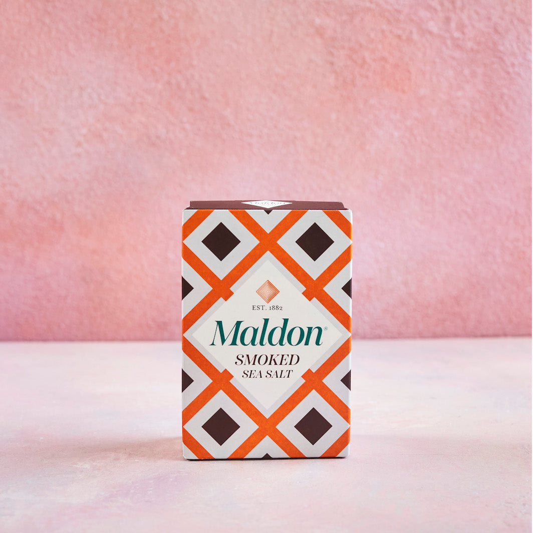 A box of Maldon Smoked Sea Salt standing upright on a light pink surface against a textured pink background. The packaging features a white base with a bold orange and dark brown geometric diamond pattern and the brand name 'Maldon' in green.