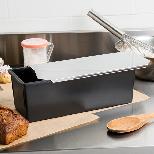 Black Matfer loaf pan with stainless steel lid on a kitchen counter next to a wooden spoon and loaf of bread.