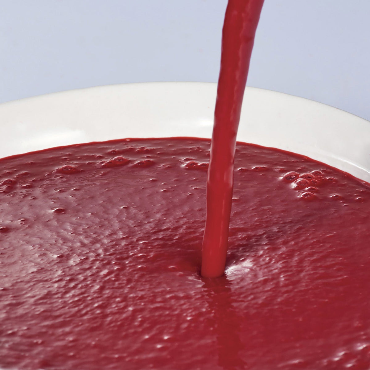 Red raspberry puree being poured into a white bowl against a light blue background