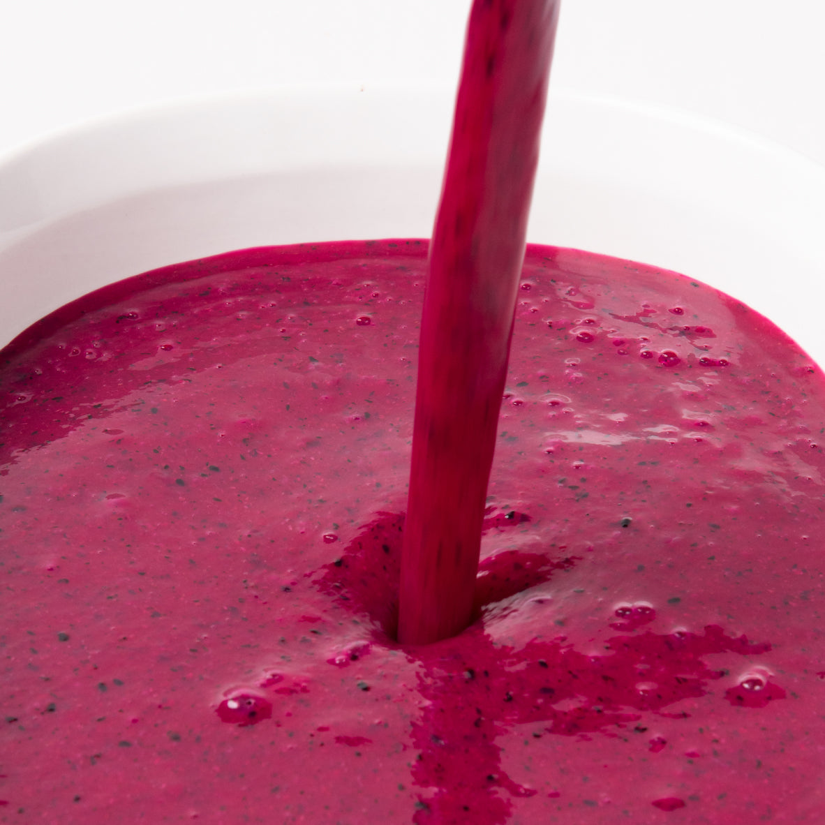 A close-up of a thick, deep magenta-red dragon fruit puree being poured from a straw into a white bowl, with visible small dark seeds throughout the drink.