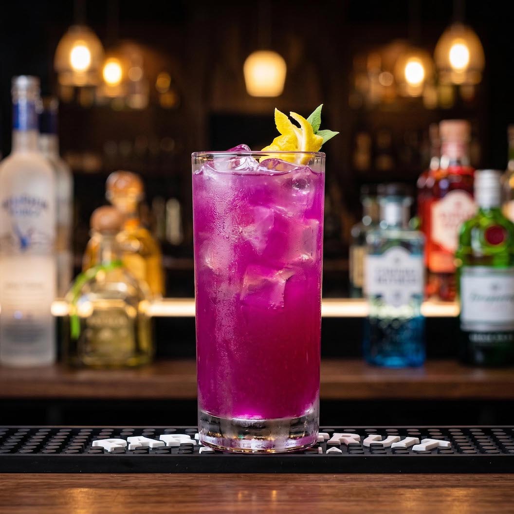 A tall glass of bright magenta-pink cocktail with ice, garnished with a yellow citrus twist, set on a bar counter with blurred liquor bottles in the background.
