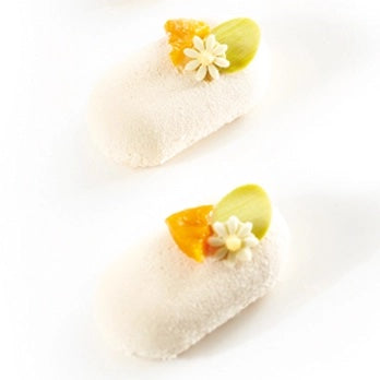 Two small white confectionery pastries decorated with a white flower, orange fruit pieces, and a green citrus slice, set against a plain white background.