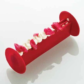 A red cylindrical yule log with circular end caps and several small pink and white flower-shaped decorations arranged along the top, photographed on a light gray background.