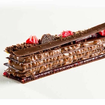 A close-up of a layered chocolate dessert or cake with glossy chocolate glaze, chocolate crumble topping, and red strawberry garnish on a white background.
