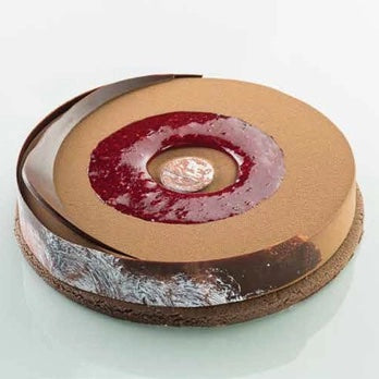 A round chocolate mousse cake with a glossy brown chocolate shell, a dark red berry compote ring, and a small gold coin in the center, set against a clean white background.