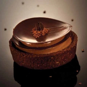 A chocolate dessert tart with a glossy dark chocolate top, a small coffee bean decoration, and a textured brown base, photographed on a dark reflective surface against a warm gradient background.