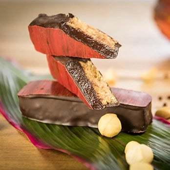 Chocolate-covered snack bars stacked on a green leaf with hazelnuts, photographed on a wooden surface with a softly blurred background.