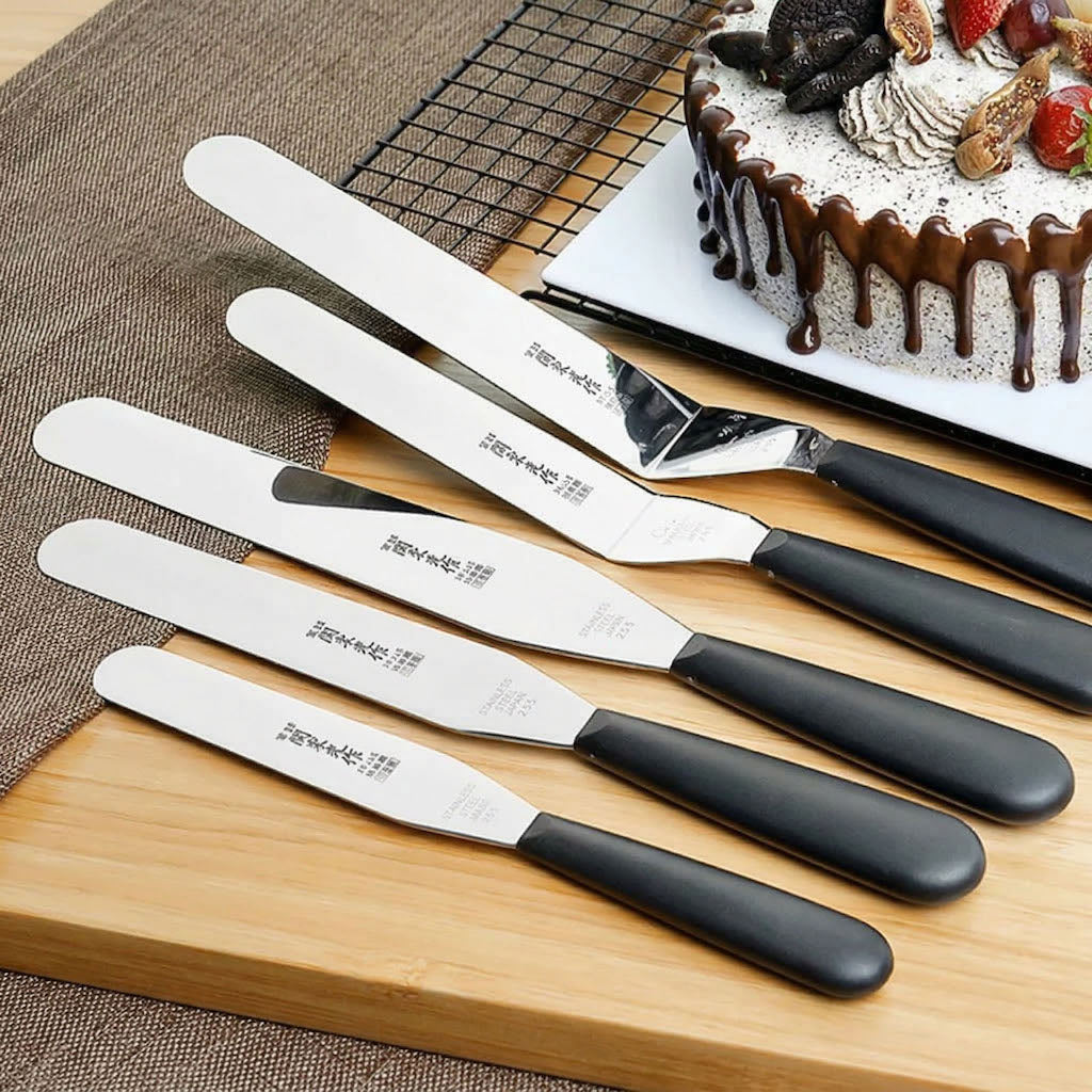 Set of five Sanneng spatulas with black handles on a wooden board, with a cake in the background.