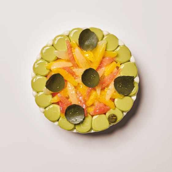 A round decorative dessert or tart topped with pale green slices around the edge, yellow-orange fruit pieces in the center, and several dark green matcha inspiration chocolate decorations, photographed from above on a light neutral background.