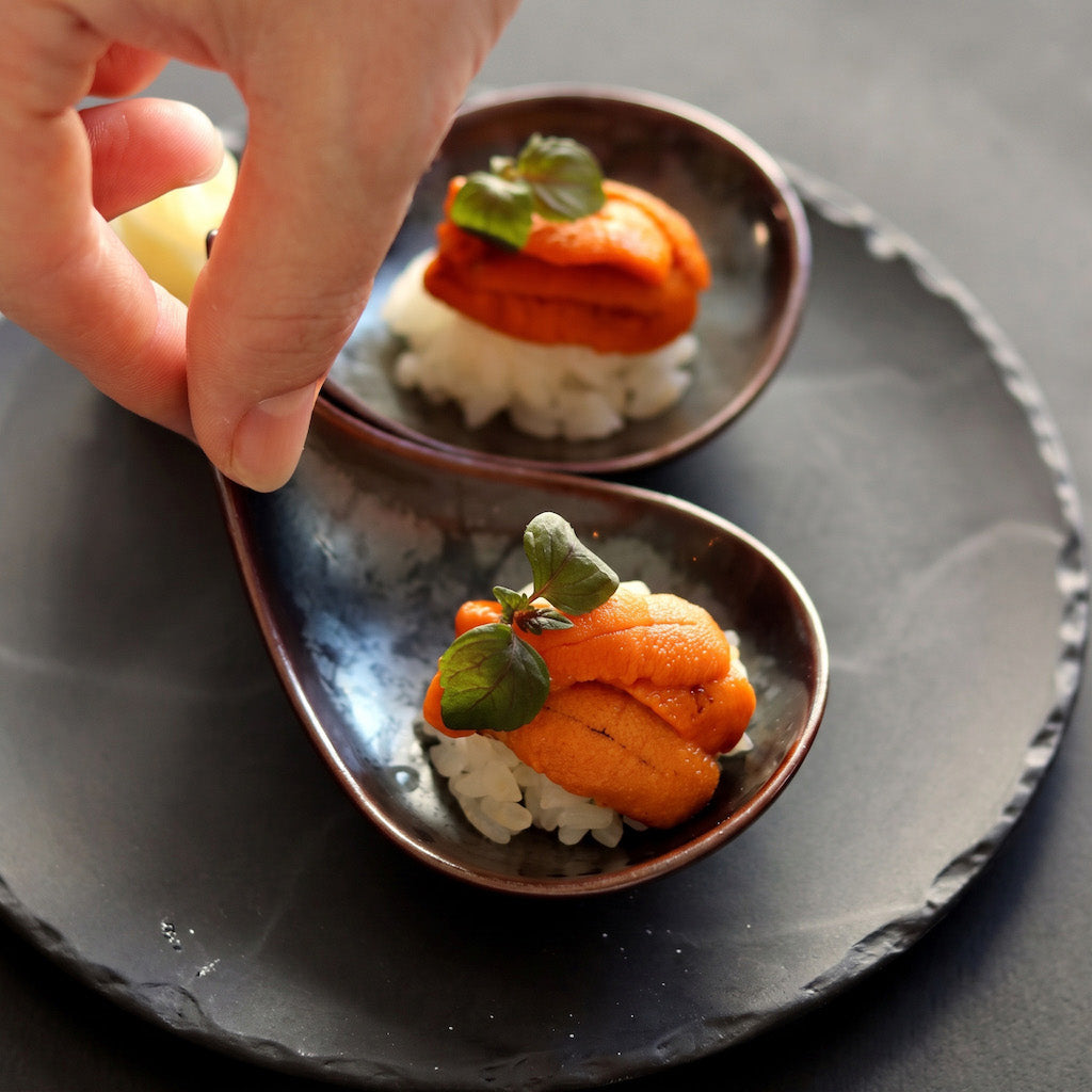Two small teardrop-shaped dark bowls on a round black plate, each holding a piece of sushi topped with orange fish and a green leaf. A hand is placing a small yellow garnish into the front bowl.