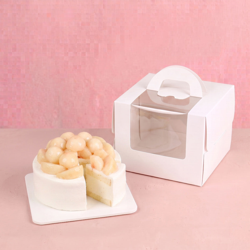 A white paper cake box with a clear window and handle sits next to a small white frosted cake with a slice cut out on a pink background.