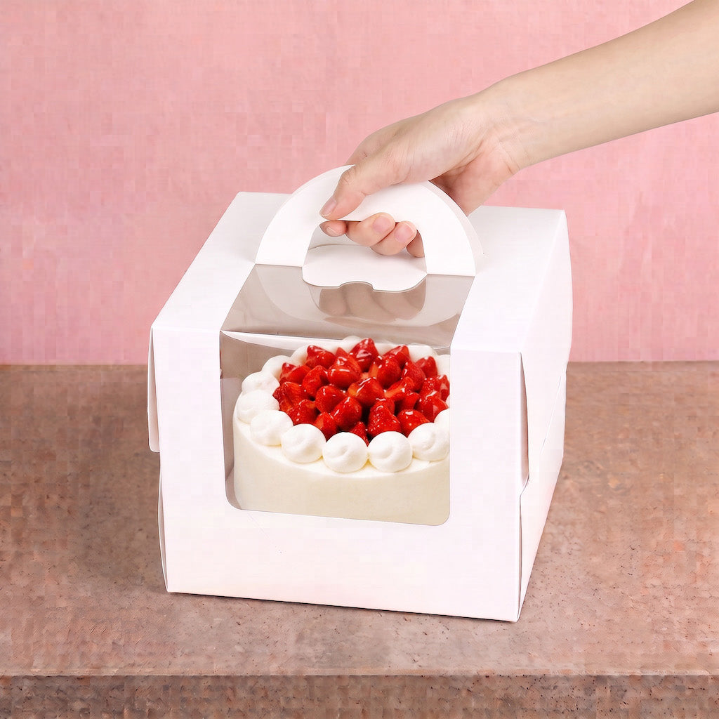 A white cardboard cake box with a clear window and a white handle, holding a frosted cake topped with red strawberries, with a hand lifting the handle against a pink background.