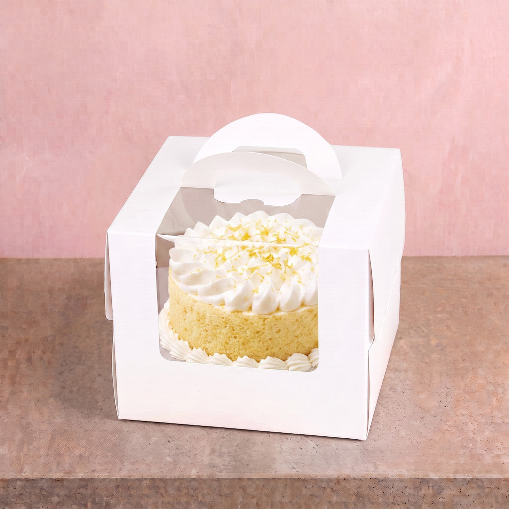 A white cardboard cake box with a clear front window and a white handle, containing a small yellow cake topped with white whipped cream and yellow sprinkles, set on a brown surface against a pink background.