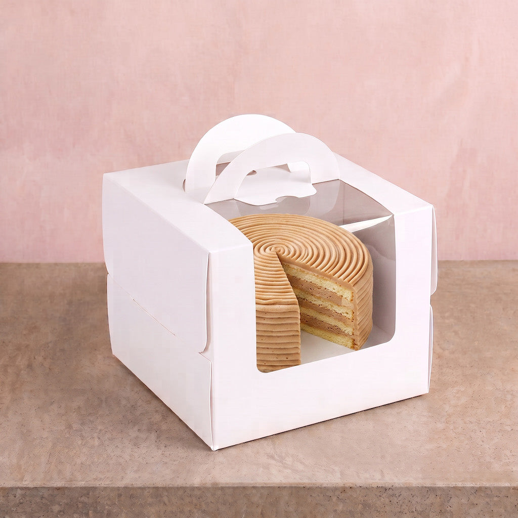 A white cardboard cake box with a clear window and paper handle, containing a layered sponge cake slice, set on a beige surface against a soft pink background.