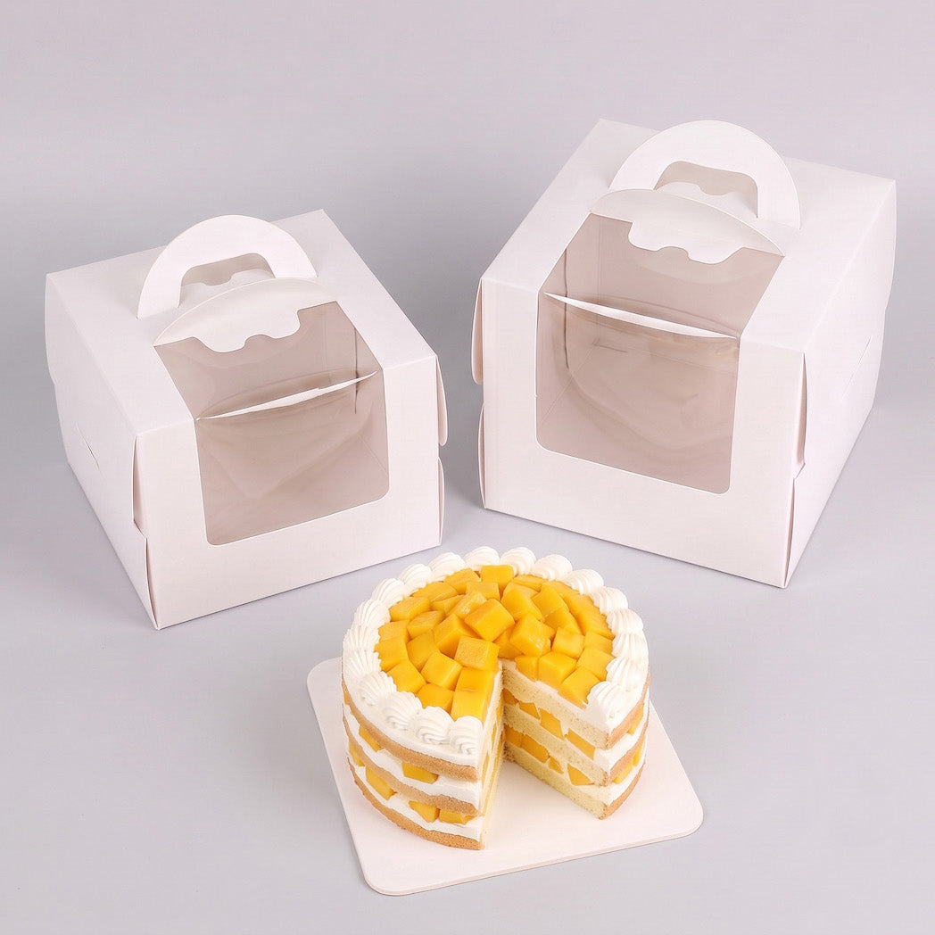 Two white cardboard cake boxes with clear window lids and built-in handles, shown on a light gray background, with a small layered mango cake placed in front.