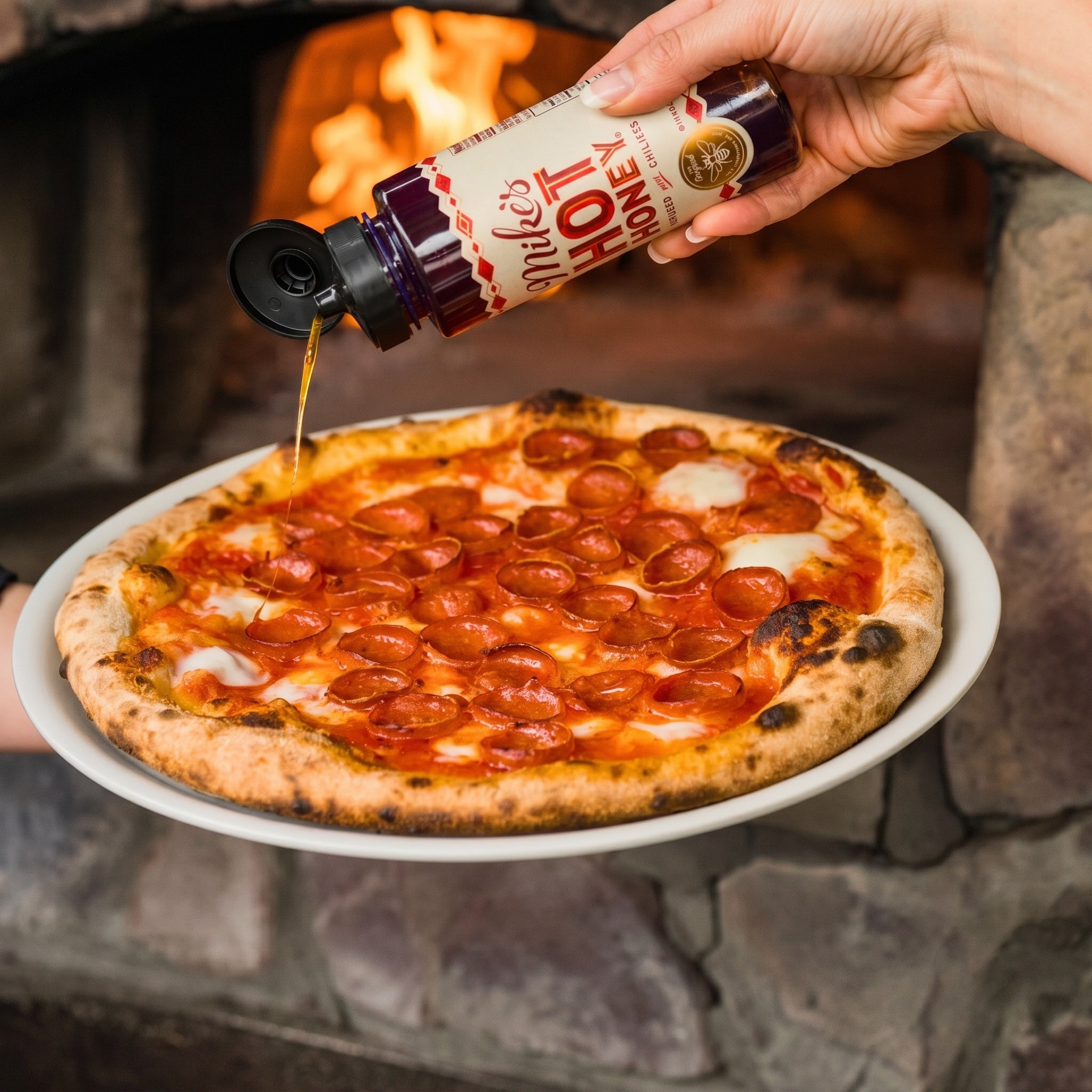 A hand pours the dark red sauce over a freshly baked pepperoni pizza on a white plate, set against a warm stone fireplace with visible flames.