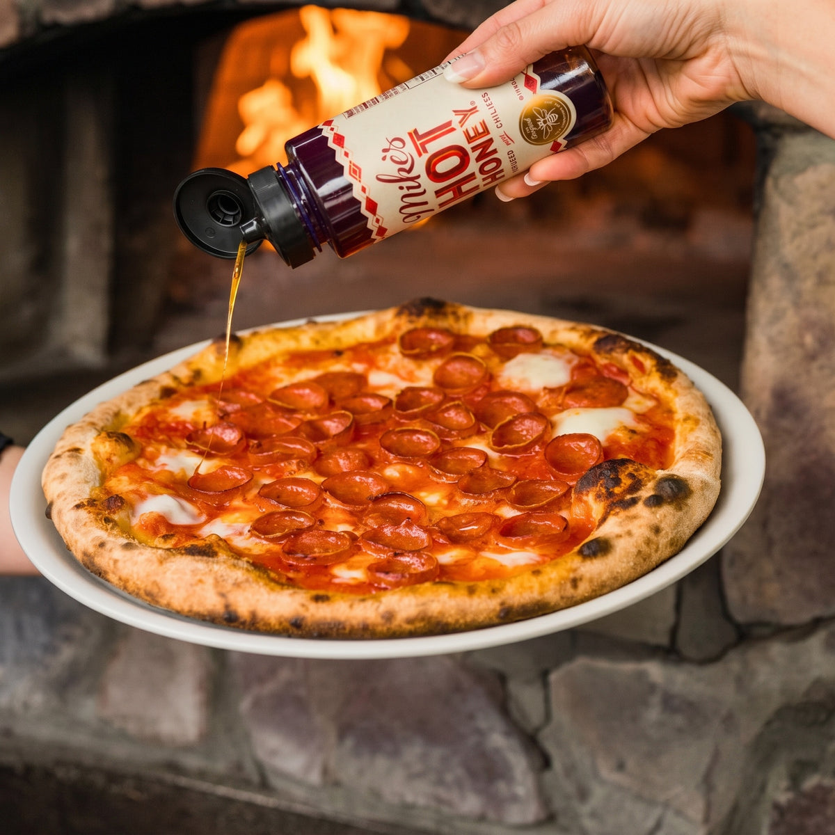 A hand pours the dark red sauce over a freshly baked pepperoni pizza on a white plate, set against a warm stone fireplace with visible flames.