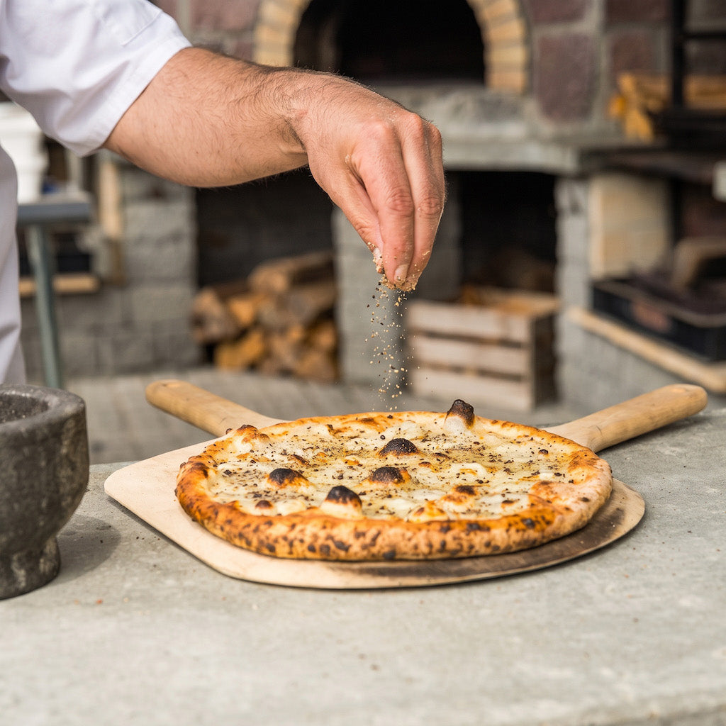 A freshly baked gluten free pizza sits on a wooden peel on a stone countertop, with a person's hand sprinkling seasoning over it. A rustic brick and stone pizza oven is blurred in the background.