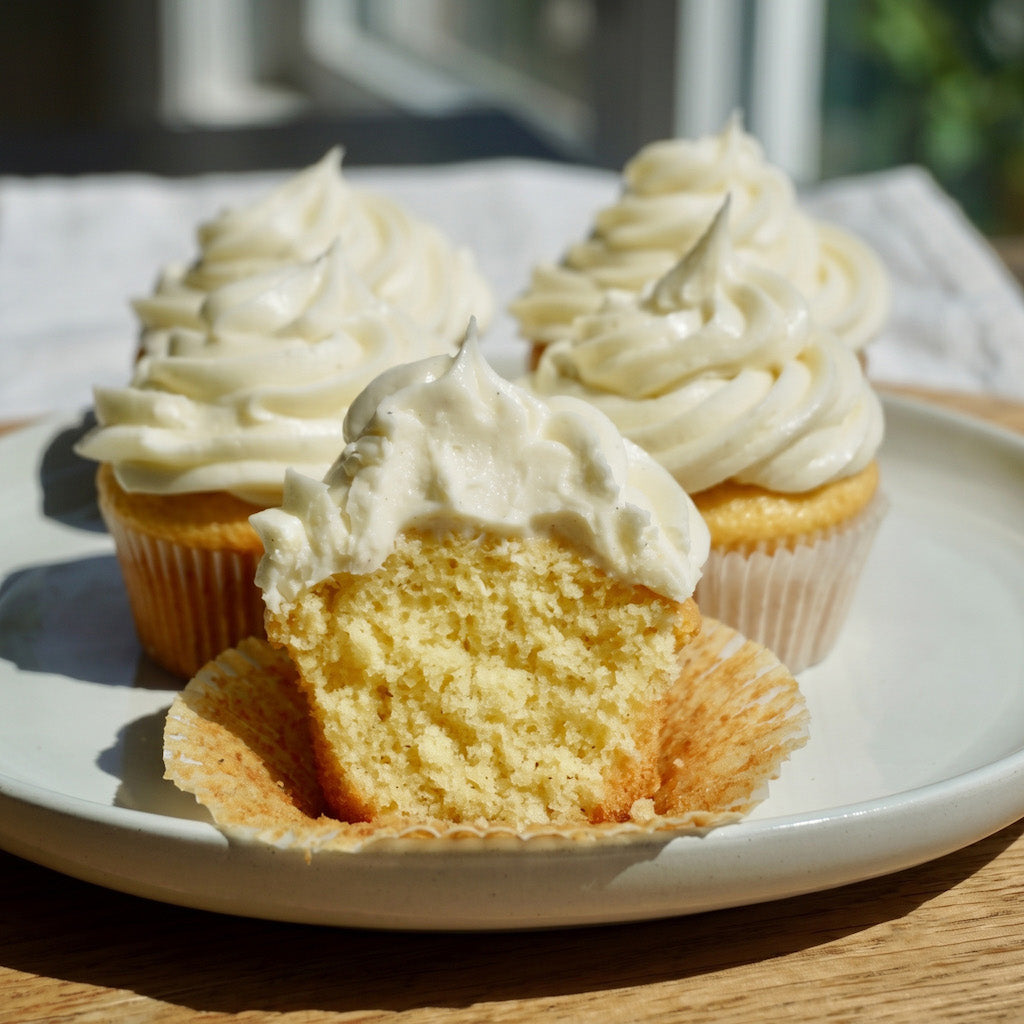 Three frosted gluten free cupcakes on a white plate, with one cupcake broken open in front to show the cake crumb. The cupcakes have thick white frosting over a light yellow cake, set on a wooden surface with a blurred indoor background.