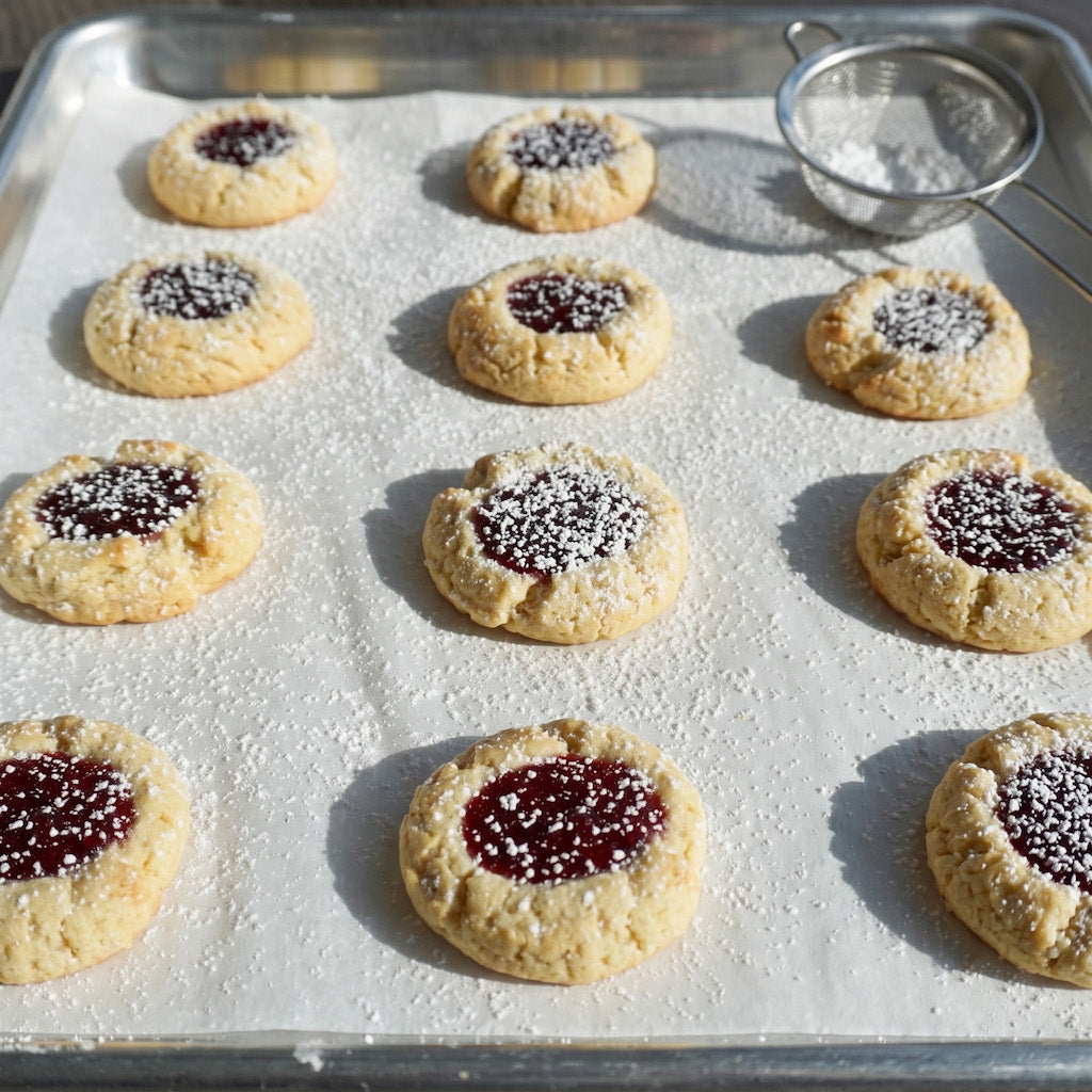 A baking sheet filled with gluten free thumbprint cookies, some topped with red jam centers and others with white powdered sugar sprinkles, set on a white parchment-lined tray with a small metal sifter in the corner.