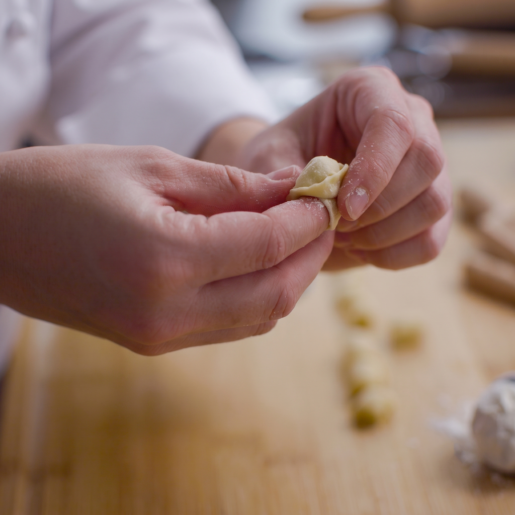 Close-up of a person's hands folding a small dumpling on a wooden cutting board, with a white chef's coat blurred in the background.