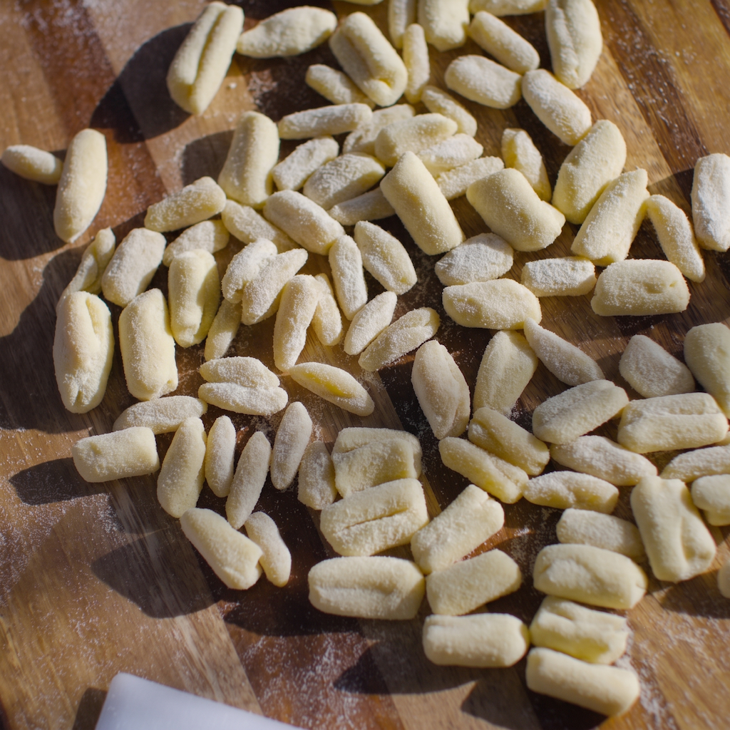 Small uncooked pasta gnocchi pieces scattered on a wooden cutting board with visible flour dusting.