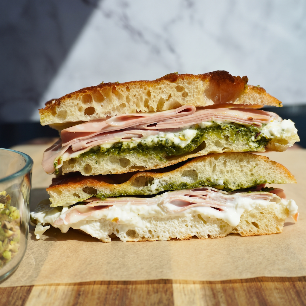 A close-up of a sandwich cut in half, showing layers of ham, white spread, and green pesto on ciabatta bread, placed on parchment paper on a wooden surface.