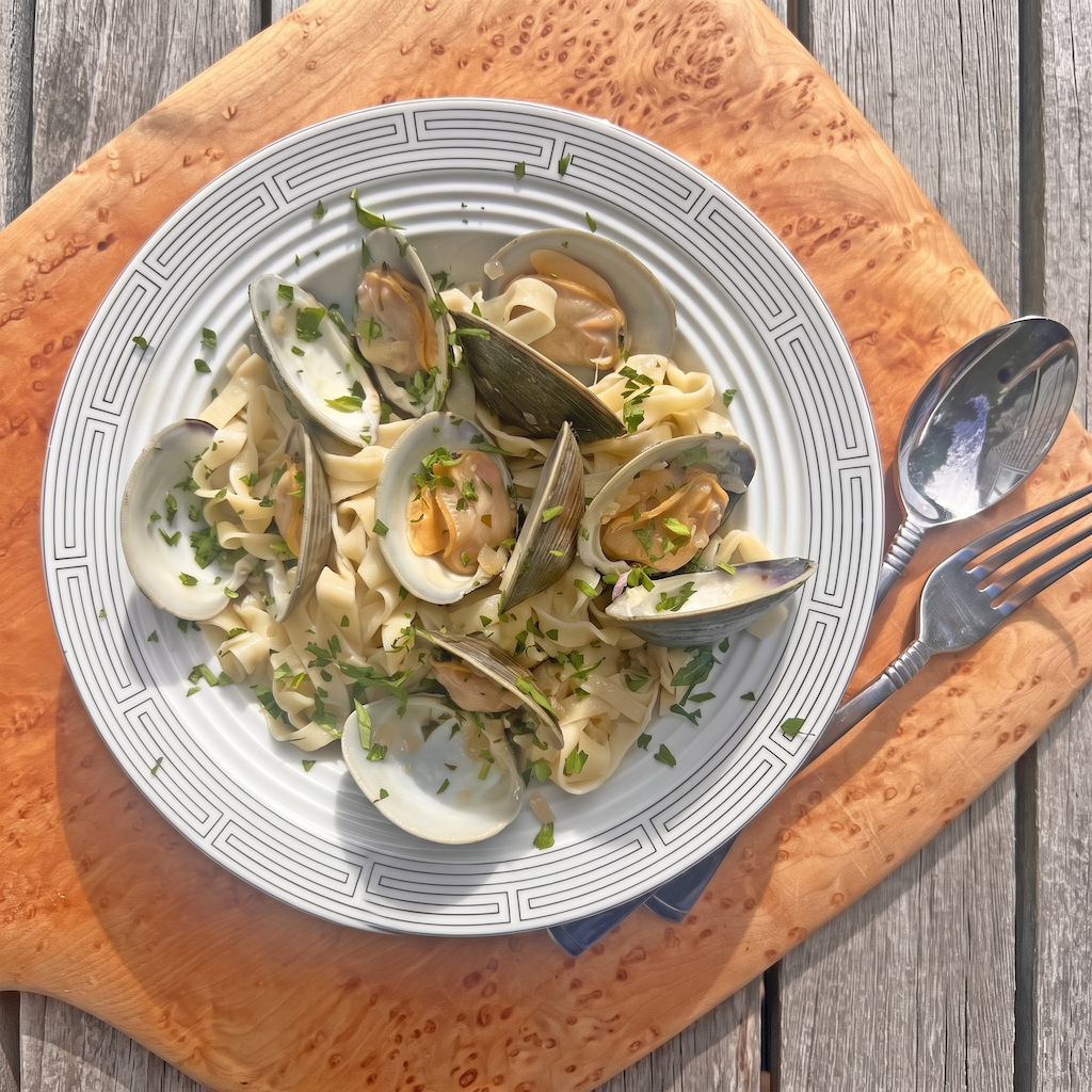A top-down view of a seafood pasta dish served on a white patterned plate, placed on a wooden board over a gray wooden table. The pasta contains mussels, shrimp, and green herbs.