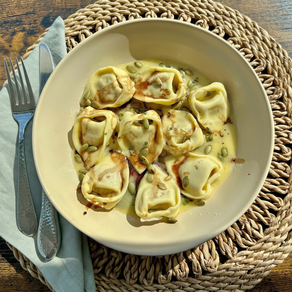 A bowl of stuffed pasta with a creamy sauce, topped with pumpkin seeds, served on a woven placemat with a fork and knife on a wooden table.